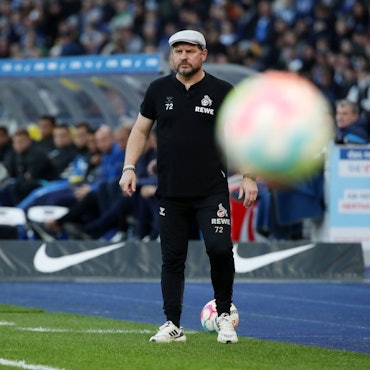 Trainer Steffen Baumgart steht während der Partie des 1. FC Köln bei Hertha BSC wie versteinert am Spielfeldrand im Berliner Olympiastadion, im Vordergrund fliegt ein Ball durchs Bild.