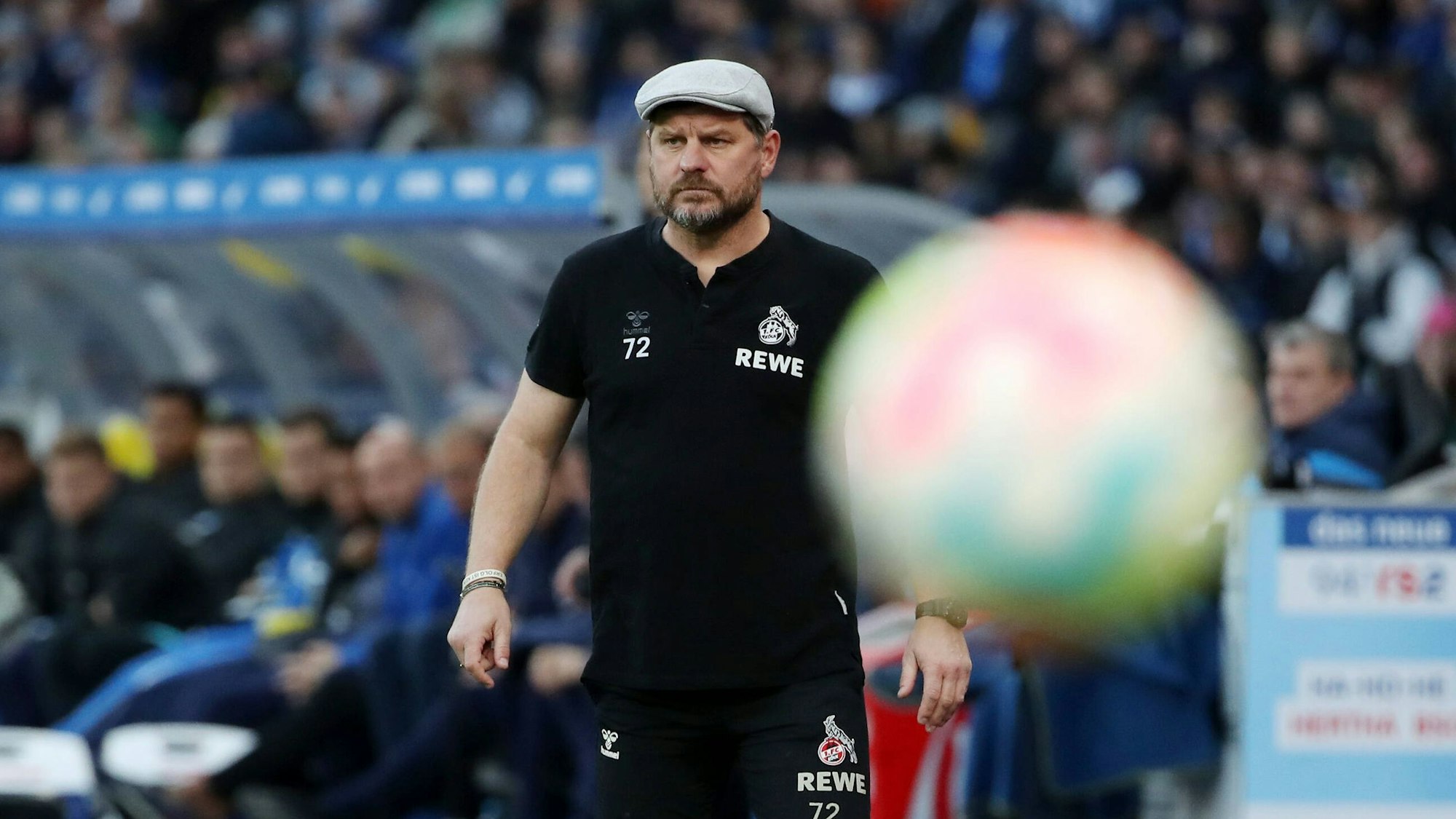 Trainer Steffen Baumgart steht während der Partie des 1. FC Köln bei Hertha BSC wie versteinert am Spielfeldrand im Berliner Olympiastadion, im Vordergrund fliegt ein Ball durchs Bild.