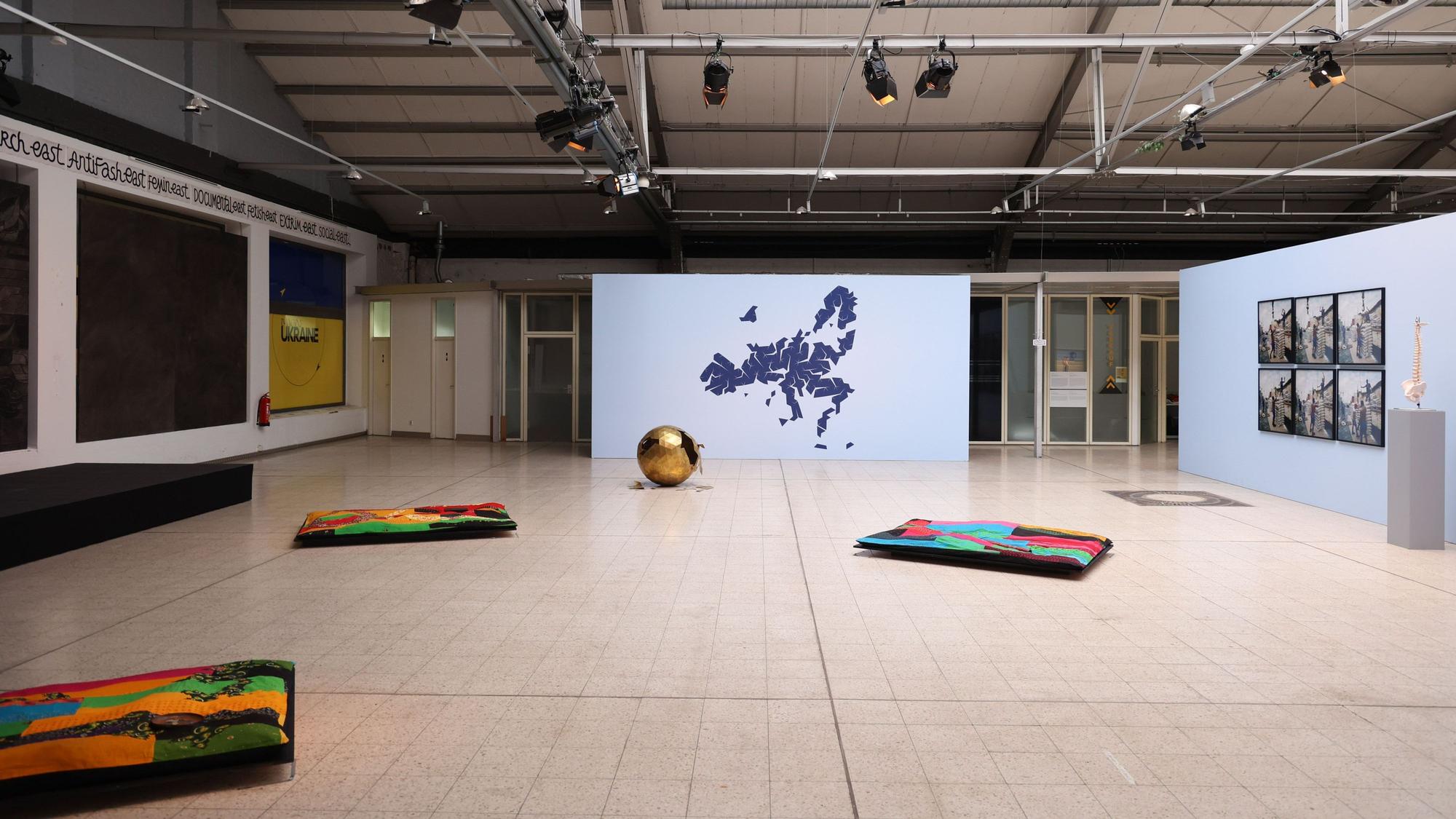 Eine Industriehalle mit verschiedenen Kunstwerken, etwa Teppichen und einer stilisierten Landkarte.