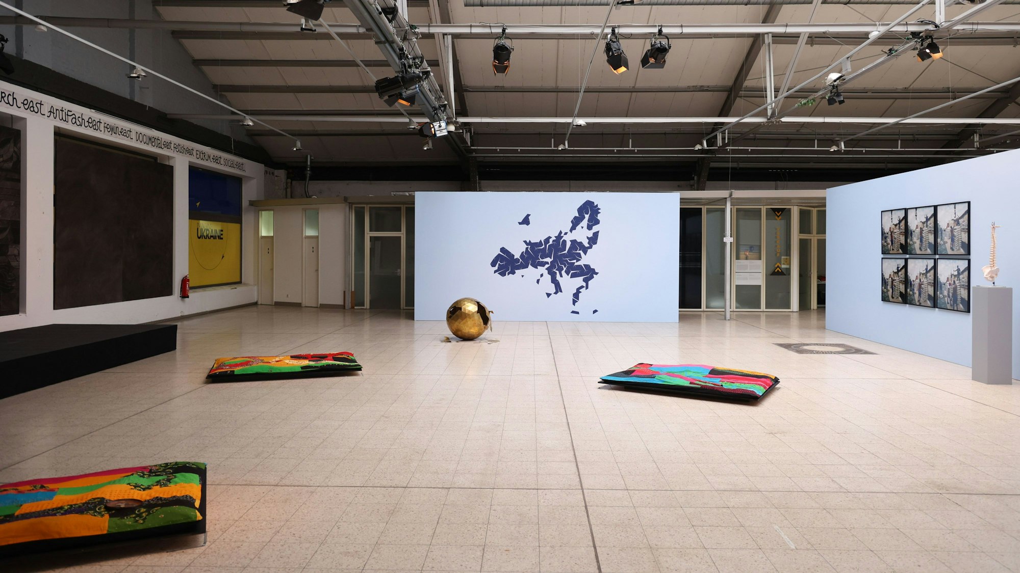 Eine Industriehalle mit verschiedenen Kunstwerken, etwa Teppichen und einer stilisierten Landkarte.