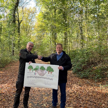 Forstamtsleiter Stephan Schütte und Uwe Zaar, Kreisdezernent Umwelt, Planung und Verkehr, halten im Königsdorfer Forst den Vertrag hoch, mit dem die Zusammenarbeit besiegelt wurde.