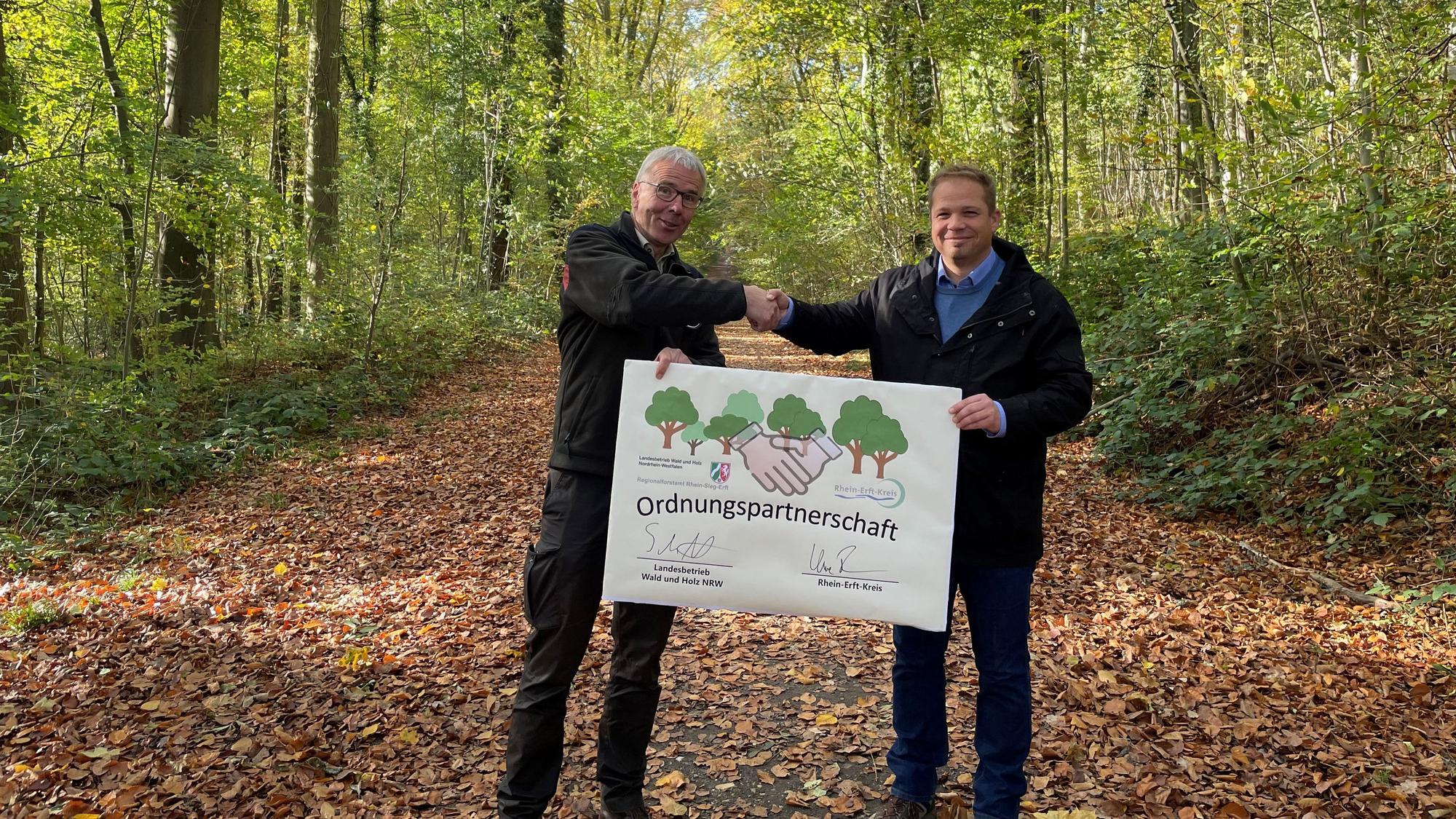 Forstamtsleiter Stephan Schütte und Uwe Zaar, Kreisdezernent Umwelt, Planung und Verkehr, halten im Königsdorfer Forst den Vertrag hoch, mit dem die Zusammenarbeit besiegelt wurde.