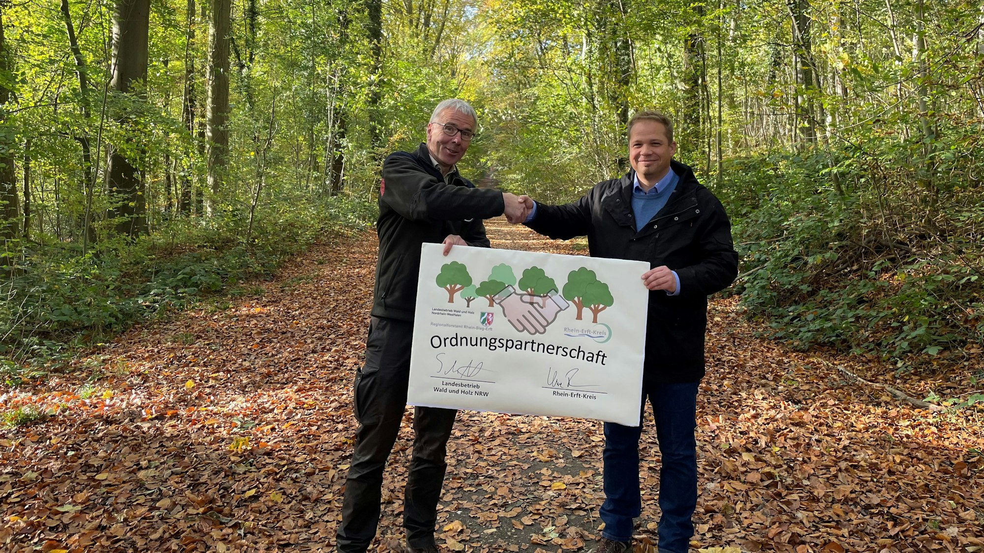 Forstamtsleiter Stephan Schütte und Uwe Zaar, Kreisdezernent Umwelt, Planung und Verkehr, halten im Königsdorfer Forst den Vertrag hoch, mit dem die Zusammenarbeit besiegelt wurde.