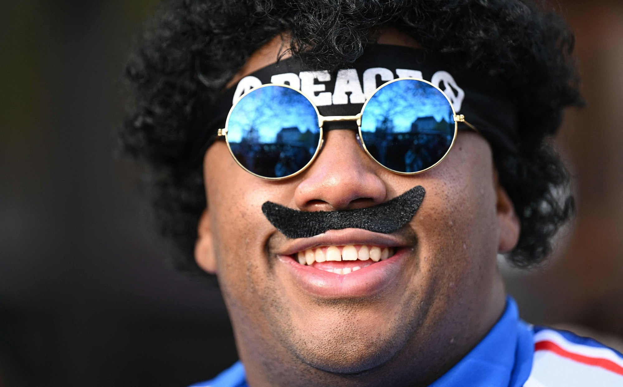 Ein Mann hat sich einen unechten Schnauzer ins Gesicht geklebt und lacht in die Kamera.