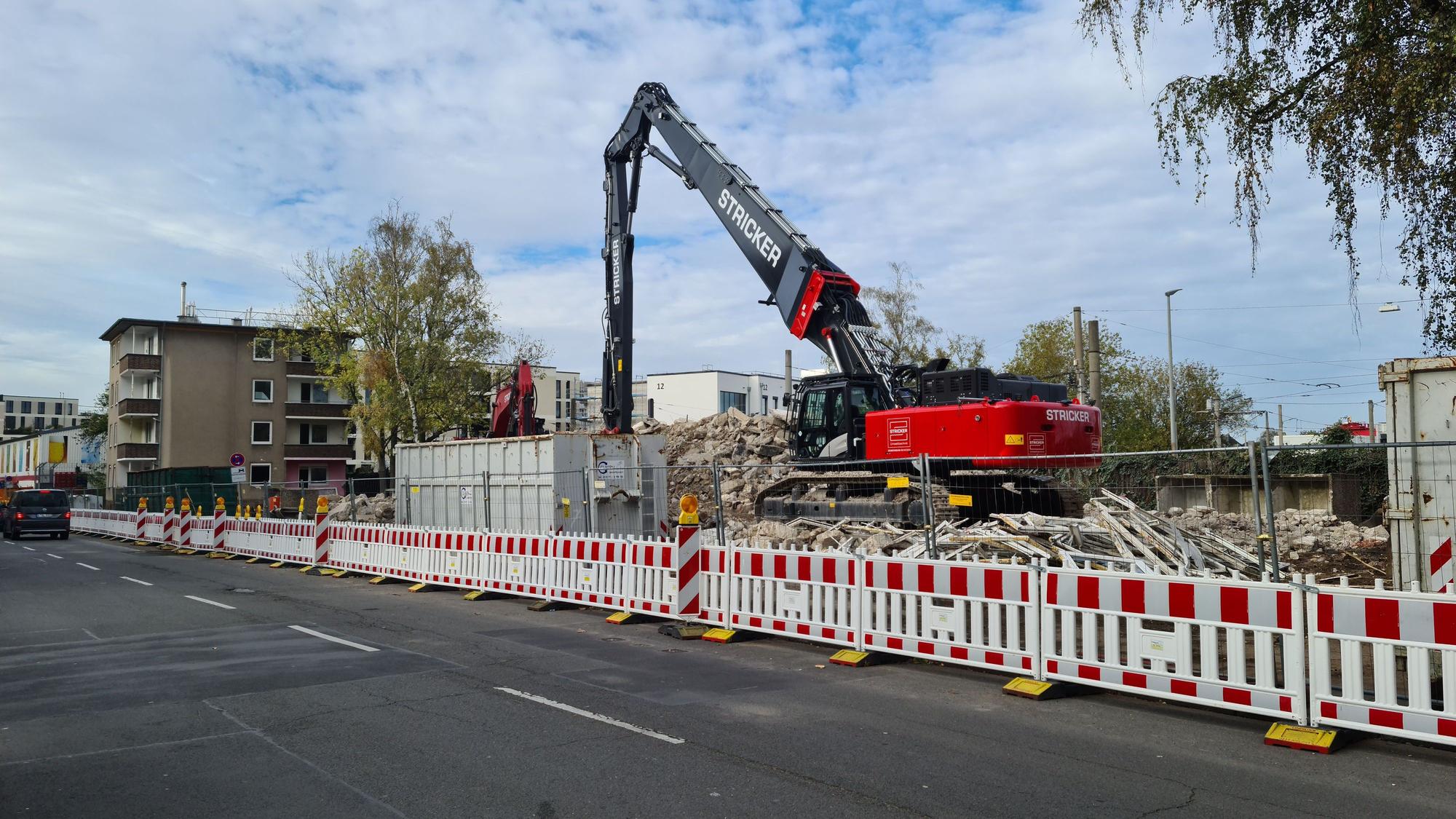 Ein Bagger steht auf einem Grundstück hinter rot-weißen Absperrungen. Es sind Trümmerteile zu sehen, aber auch noch ein Haus, das als nächstes abgerissen wird.