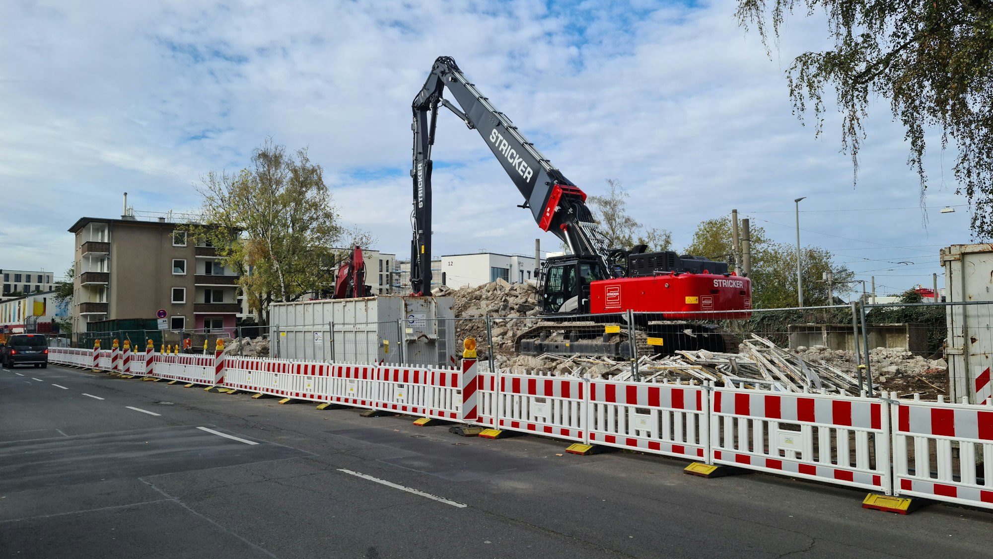 Ein Bagger steht auf einem Grundstück hinter rot-weißen Absperrungen. Es sind Trümmerteile zu sehen, aber auch noch ein Haus, das als nächstes abgerissen wird.