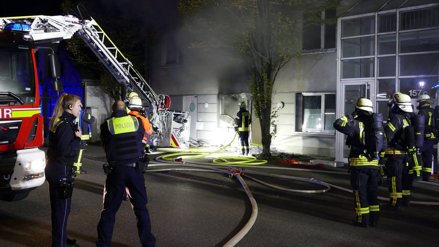 Feuerwehrleute stehen in der Nacht vor einem Fenster, aus dem dicker, schwarzer Rauch quillt. Ein Feuerwehrwagen steht daneben, ein Schlauch liegt auf der Straße.
