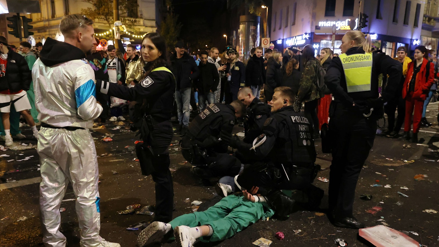 Zwei Polizisten knien auf der Zülpicher Straße und fesseln einem Mann, der auf dem Bauch liegt, die Hände auf dem Rücken zusammen. Zwei Mitarbeiterinnen des Ordnungsamtes stehen daneben und halten Schaulustige fern.