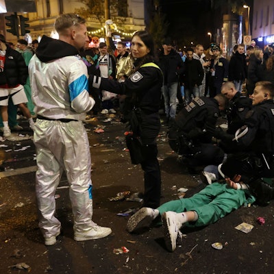 Zwei Polizisten knien auf der Zülpicher Straße und fesseln einem Mann, der auf dem Bauch liegt, die Hände auf dem Rücken zusammen. Zwei Mitarbeiterinnen des Ordnungsamtes stehen daneben und halten Schaulustige fern.