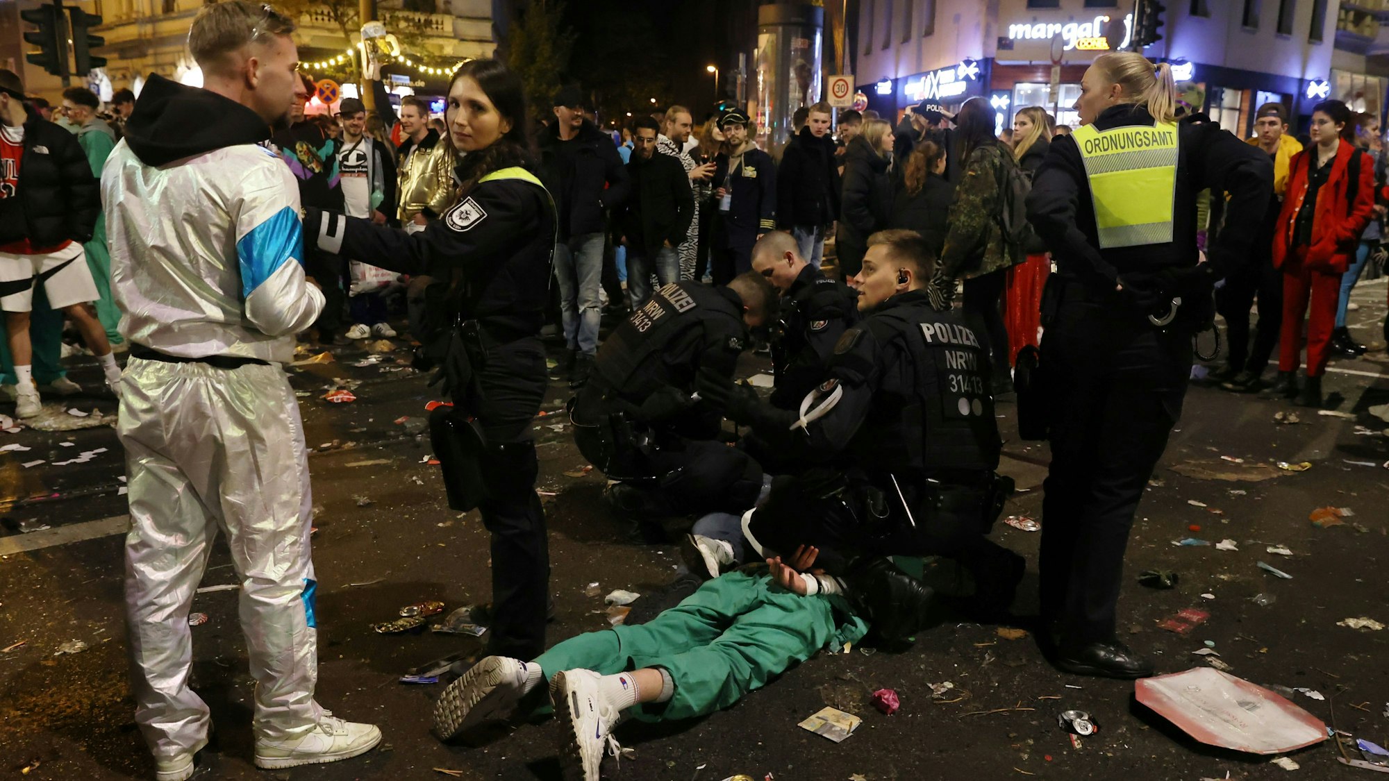Zwei Polizisten knien auf der Zülpicher Straße und fesseln einem Mann, der auf dem Bauch liegt, die Hände auf dem Rücken zusammen. Zwei Mitarbeiterinnen des Ordnungsamtes stehen daneben und halten Schaulustige fern.
