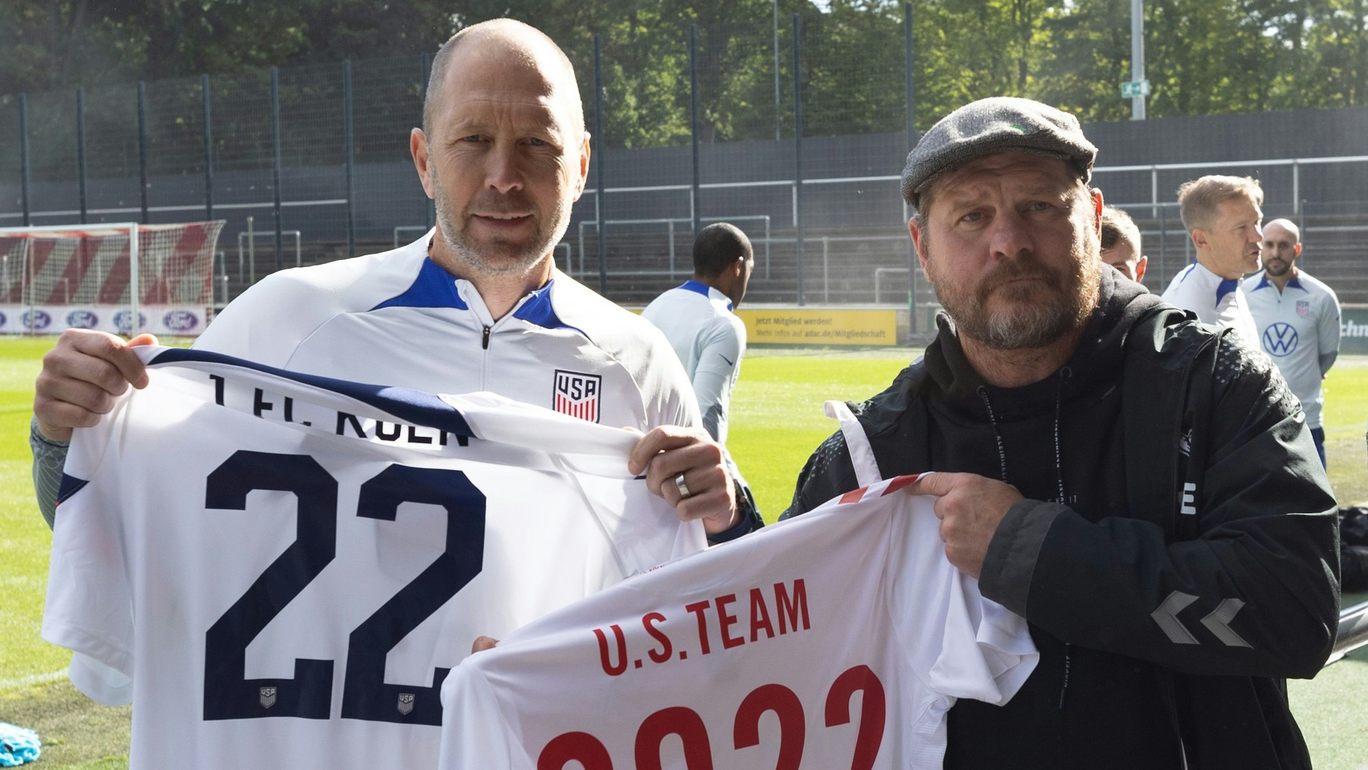 20.09.2022 in Köln: US-Nationaltrainer Gregg Berhalter trifft Kölns Trainer im Franz-Kremer-Stadion. Beide halten Trikots in der Hand, Baumgart trägt eine Mütze, Berhalter hat ein weißes Trainingsshirt an.