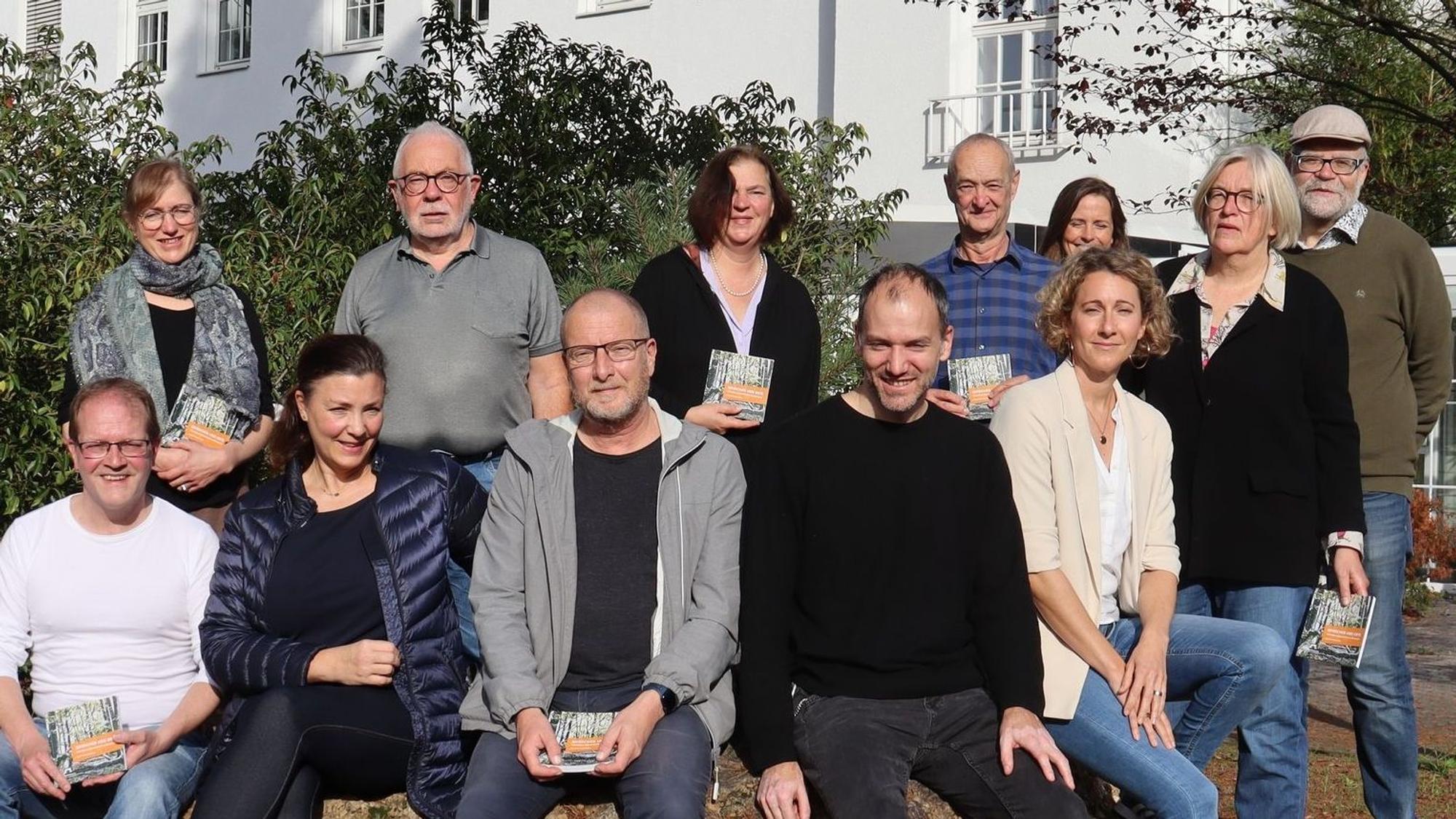 Ingo Müller-Becker (vorn l.) lud Dellbrücker Kulturschaffende ein, die jeweils eigene Sicht auf ihr Veedel auszudrücken. Zwölf Menschen sitzen und stehen in zwei Reihen vor einem weißen Gebäude.