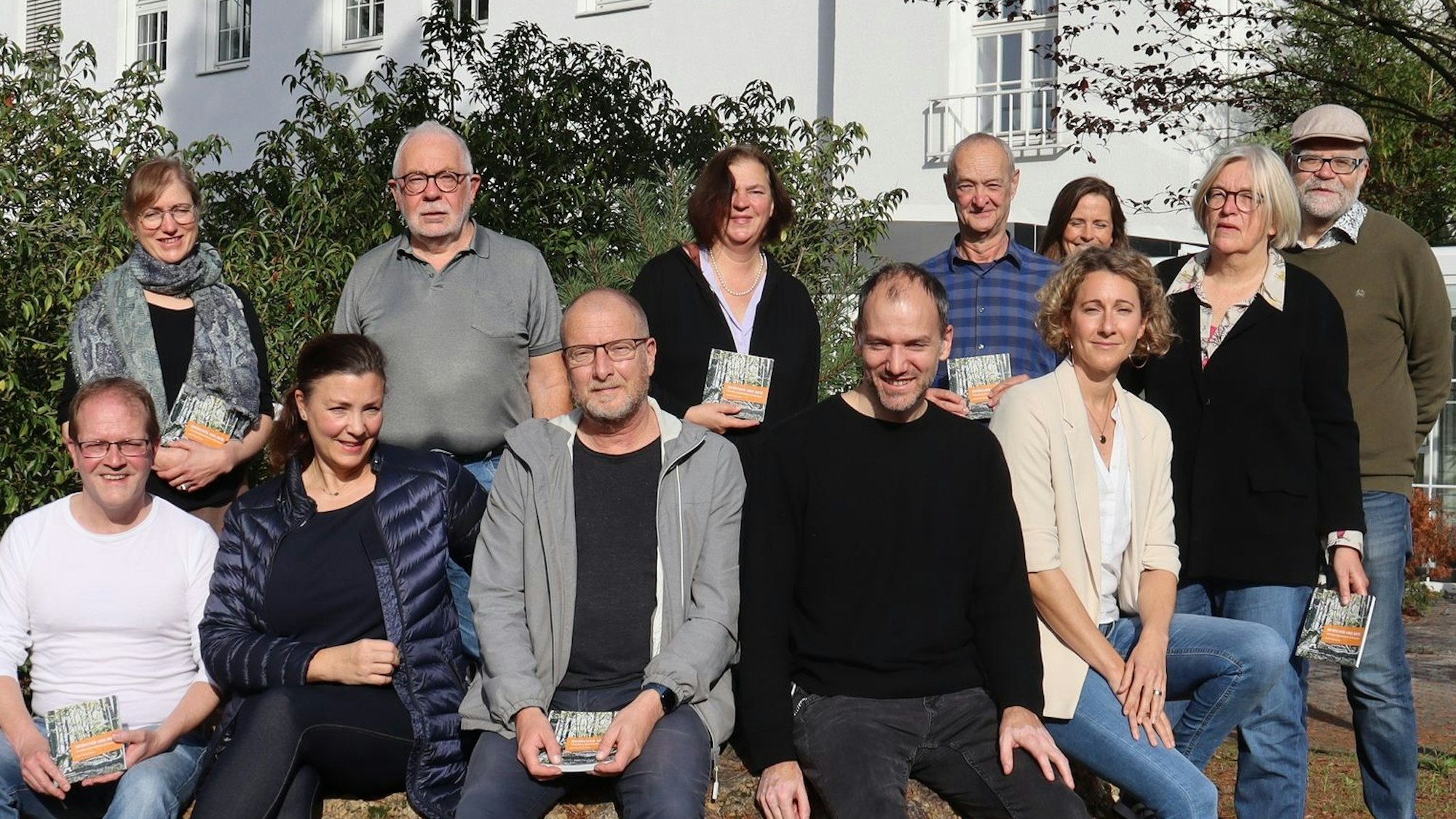 Ingo Müller-Becker (vorn l.) lud Dellbrücker Kulturschaffende ein, die jeweils eigene Sicht auf ihr Veedel auszudrücken. Zwölf Menschen sitzen und stehen in zwei Reihen vor einem weißen Gebäude.
