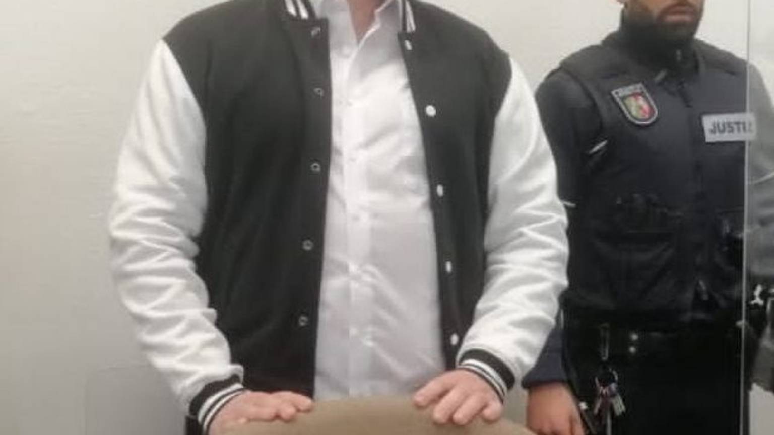 Der Angeklagte steht hinter einem Stuhl im Gerichtssaal. Er trägt eine schwarz-weiße Jacke, neben ihm steht ein Justiz-Beamter. Das Gesicht des Angeklagten ist gepixelt.