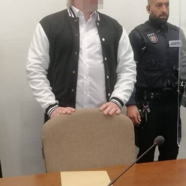 Der Angeklagte steht hinter einem Stuhl im Gerichtssaal. Er trägt eine schwarz-weiße Jacke, neben ihm steht ein Justiz-Beamter. Das Gesicht des Angeklagten ist gepixelt.