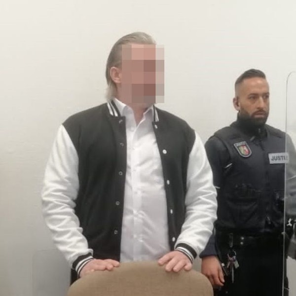Der Angeklagte steht hinter einem Stuhl im Gerichtssaal. Er trägt eine schwarz-weiße Jacke, neben ihm steht ein Justiz-Beamter. Das Gesicht des Angeklagten ist gepixelt.