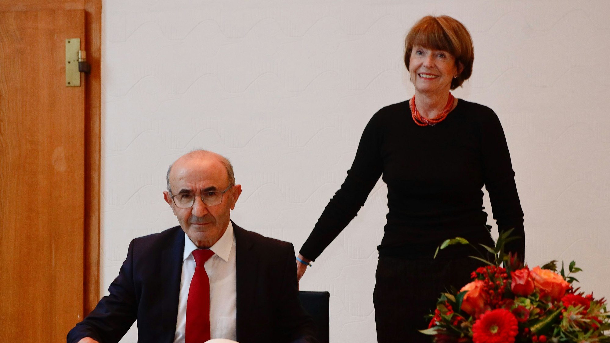 Tayfun Keltek wird zu seinem 75. Geburtstag von Kölner OB Henriette Reker im Muschelsaal empfangen. Keltek sitzt an einem Schreibtisch und trägt sich in das Gästebuch der Stadt ein. Reker steht neben ihm.