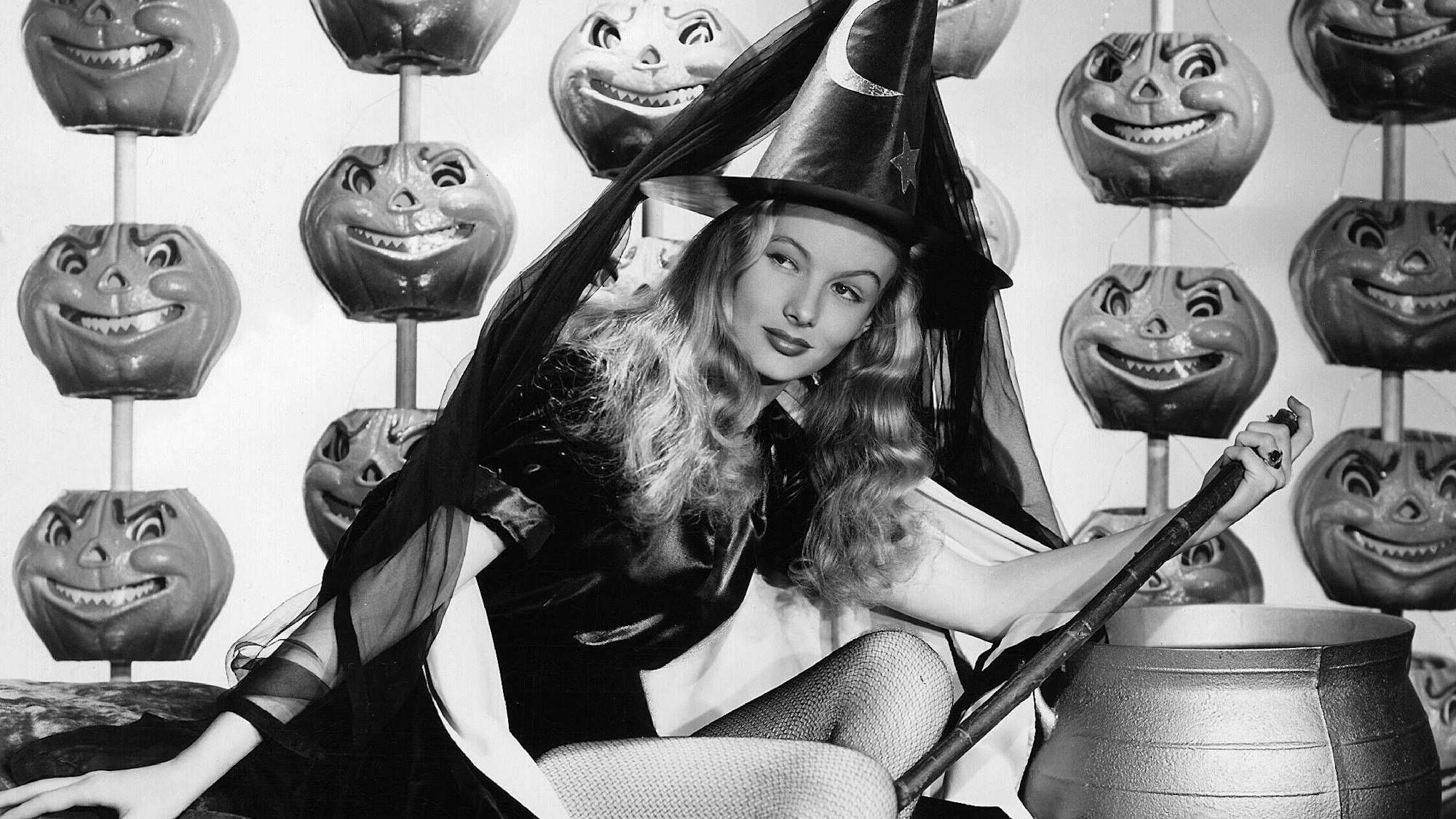 Veronica Lake als Hexe in „Meine Frau, die Hexe“. Sie sitzt mit Besen und Hexenkostüm vor einem goldenen Kessel. Im Hintergrund sind lustige Monsterkürbisse.