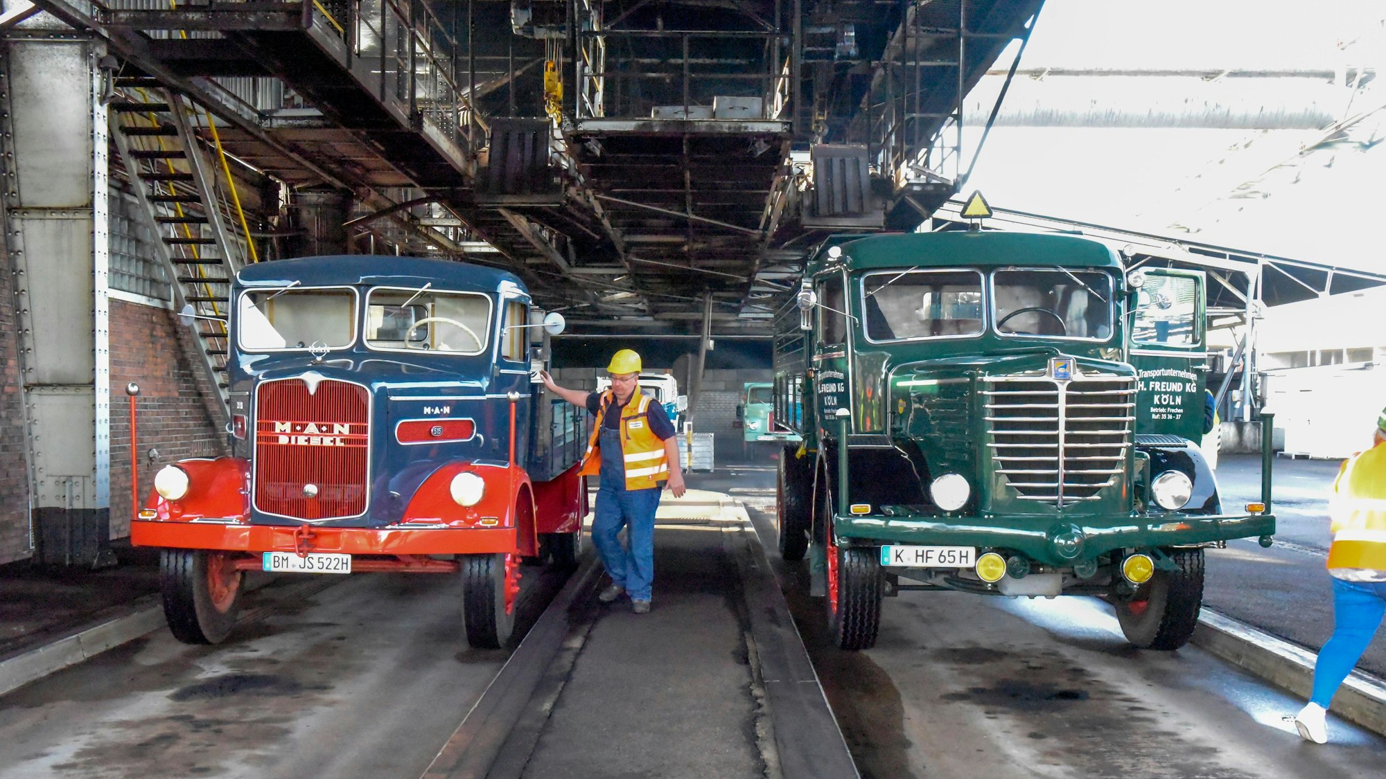 Zwei LKW aus den 1950er-Jahren stehen in der Brikettfabrik in Frechen und warten, dass sie mit Kohle beladen werden. Ein Arbeiter steht mit Helm und Warnweste neben dem linken LKW.