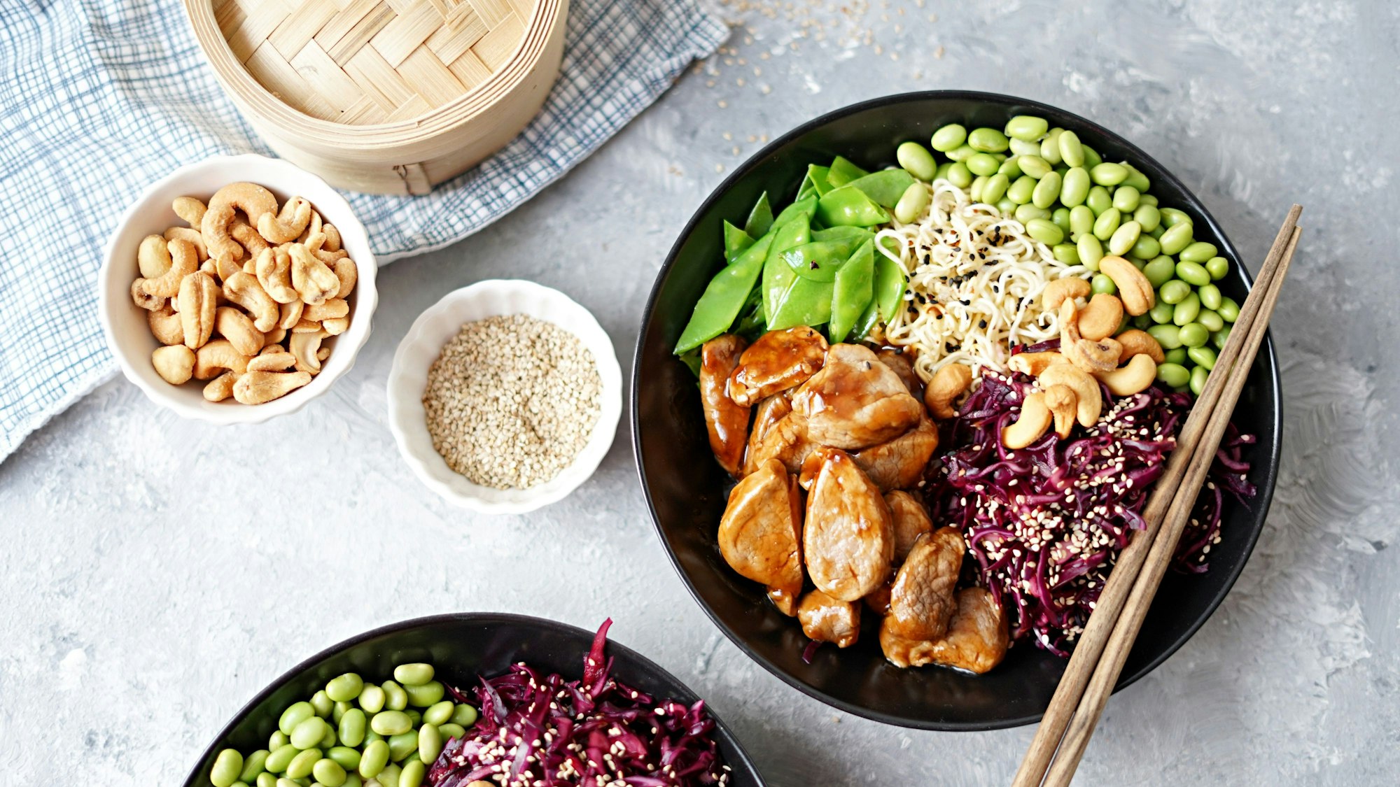 In einer schwarzen Schale sind verschiedene Zutaten nebeneinander angeordnet, daneben zwei kleinere Schalen mit Cashewkernen und Sesam