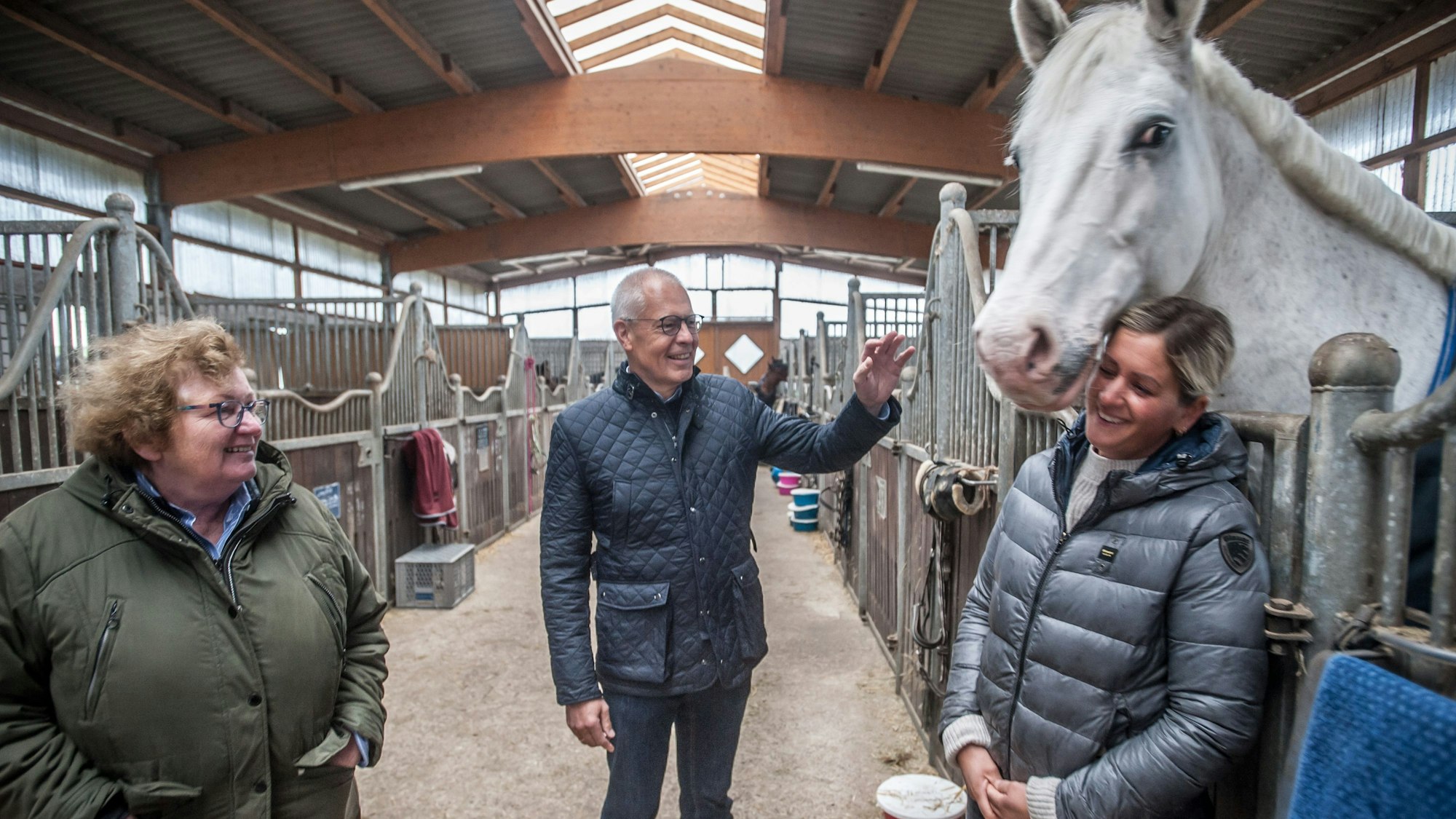 Hermann-Josef Tebroke mit Dörthe Völker (blond) und Sigrid Völker im Pferdestall vor einer Box mit einem weißen Pferd.