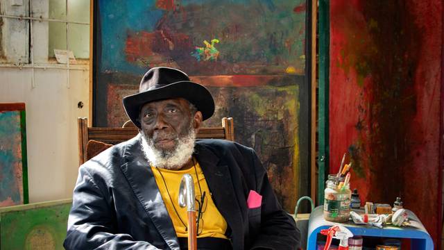Der Künstler Frank Bowling sitzt in seinem Atelier vor Leinwänden und Malutensilien.