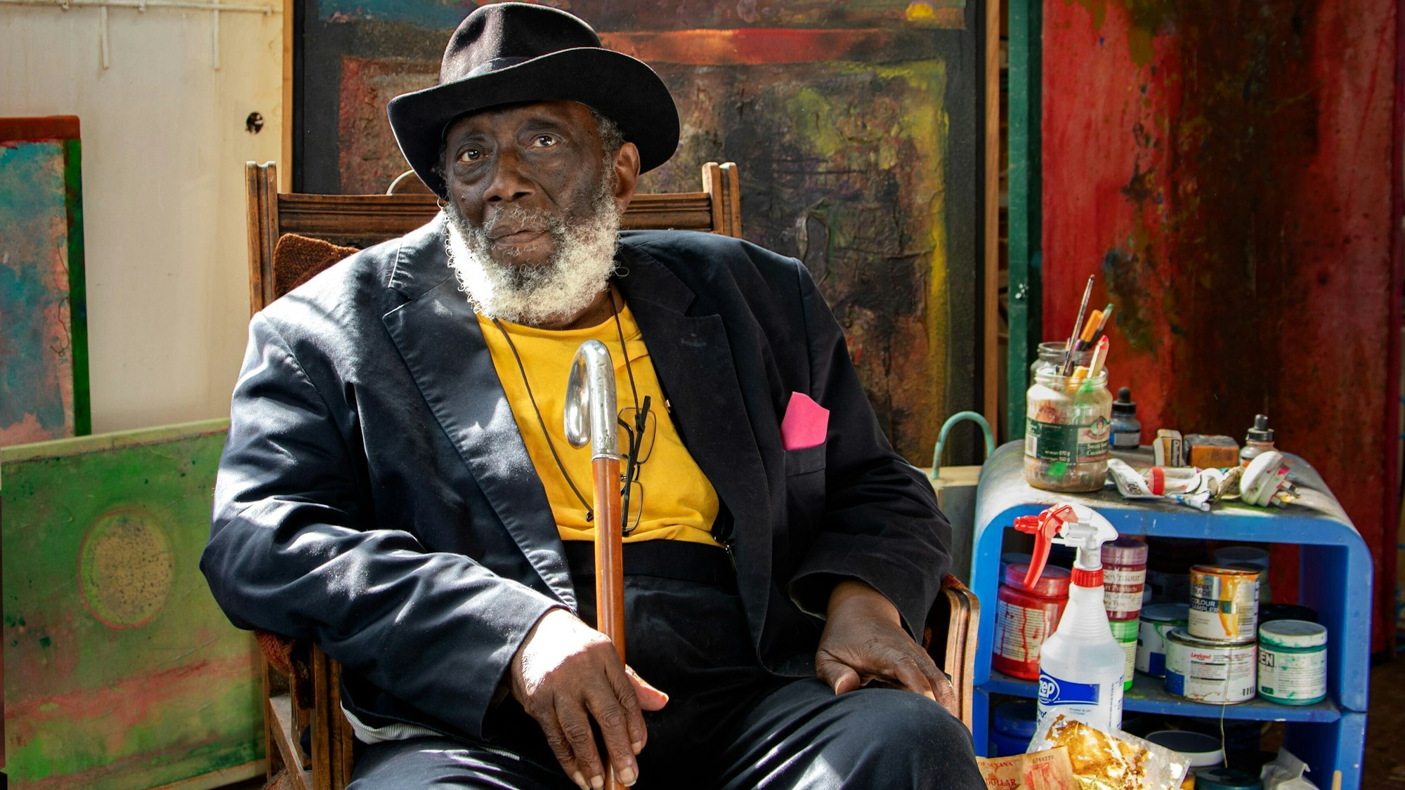 Der Künstler Frank Bowling sitzt in seinem Atelier vor Leinwänden und Malutensilien.