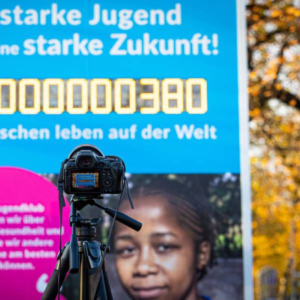 Es ist eine Kamera zu sehen, die auf die Weltbevölkerungsuhr zeigt. Die Zahl auf dieser hat acht Milliarden erreicht.