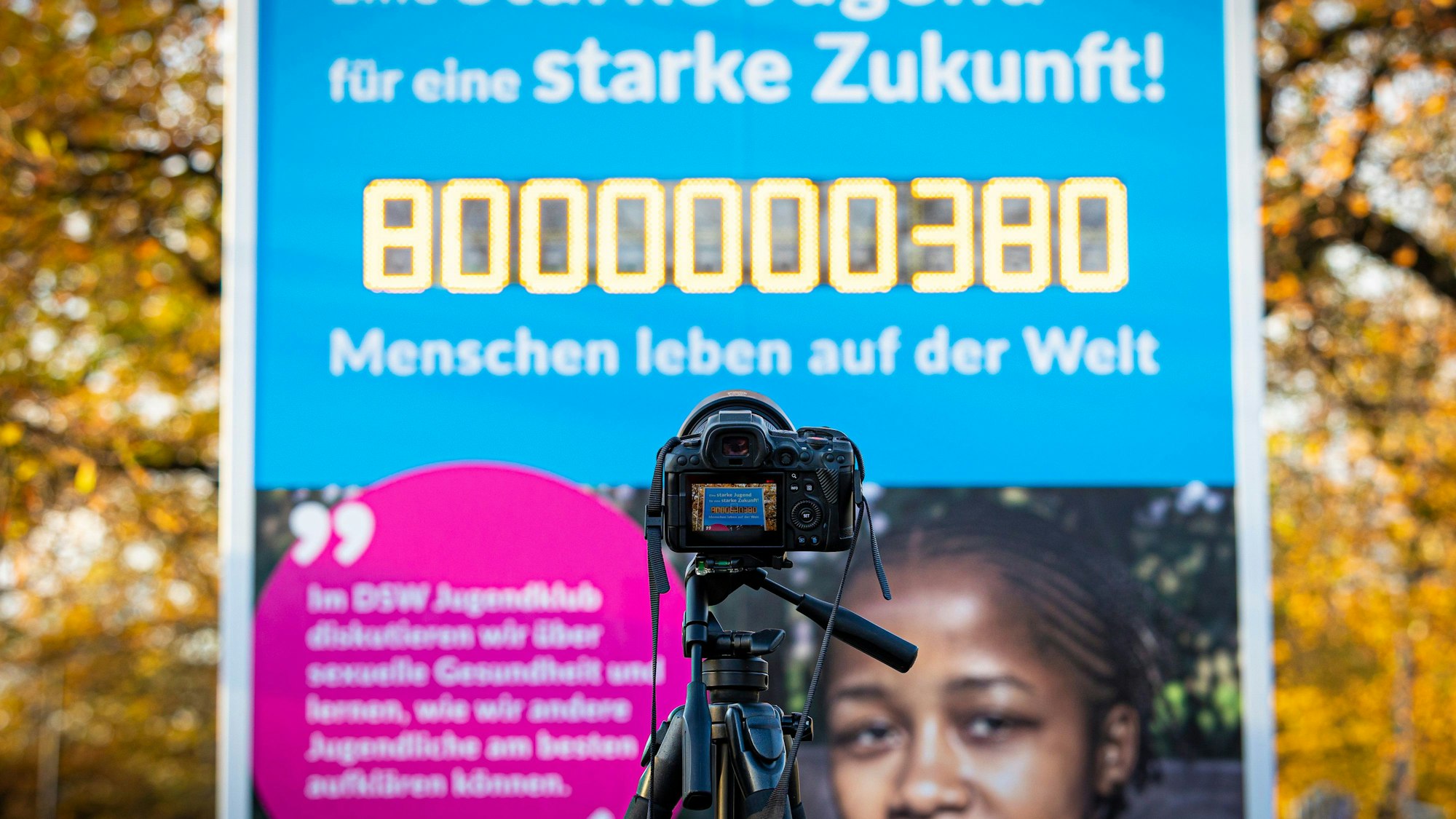 Es ist eine Kamera zu sehen, die auf die Weltbevölkerungsuhr zeigt. Die Zahl auf dieser hat acht Milliarden erreicht.