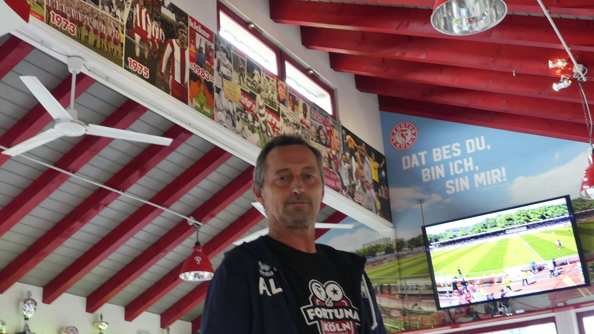 Der Wirt des Fortuna-Vereinsheims steht in seinem Gastraum vor einem Fernseher, auf dem ein Fußballspiel läuft.