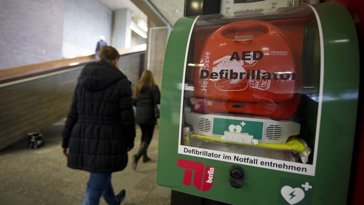 Personen gehen in einem Gebäude an einem AED-Defibrillator vorbei.