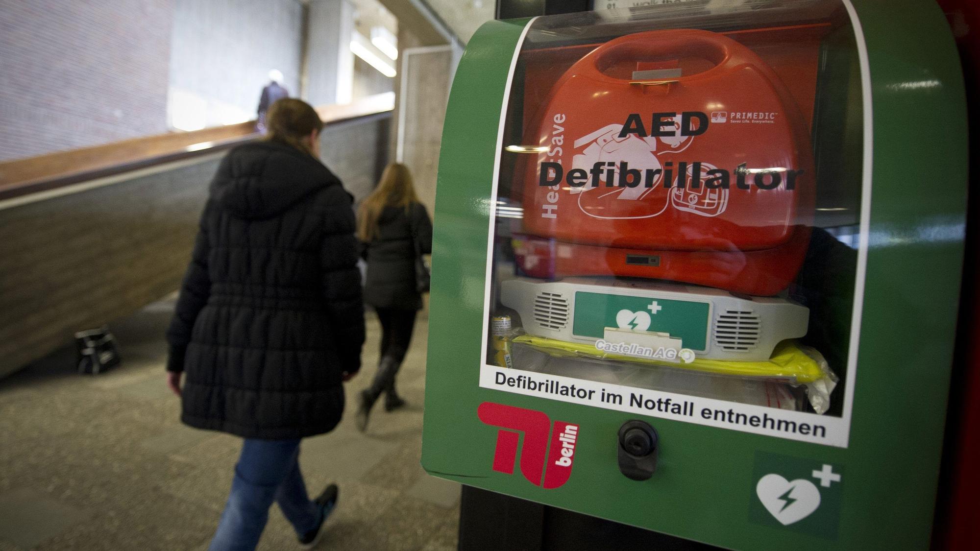 Personen gehen in einem Gebäude an einem AED-Defibrillator vorbei.