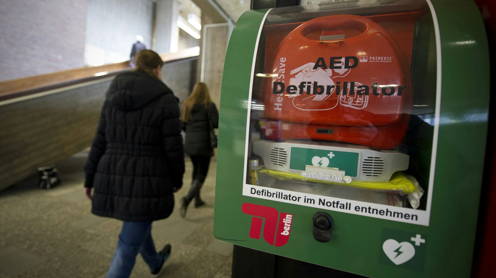 Personen gehen in einem Gebäude an einem AED-Defibrillator vorbei.