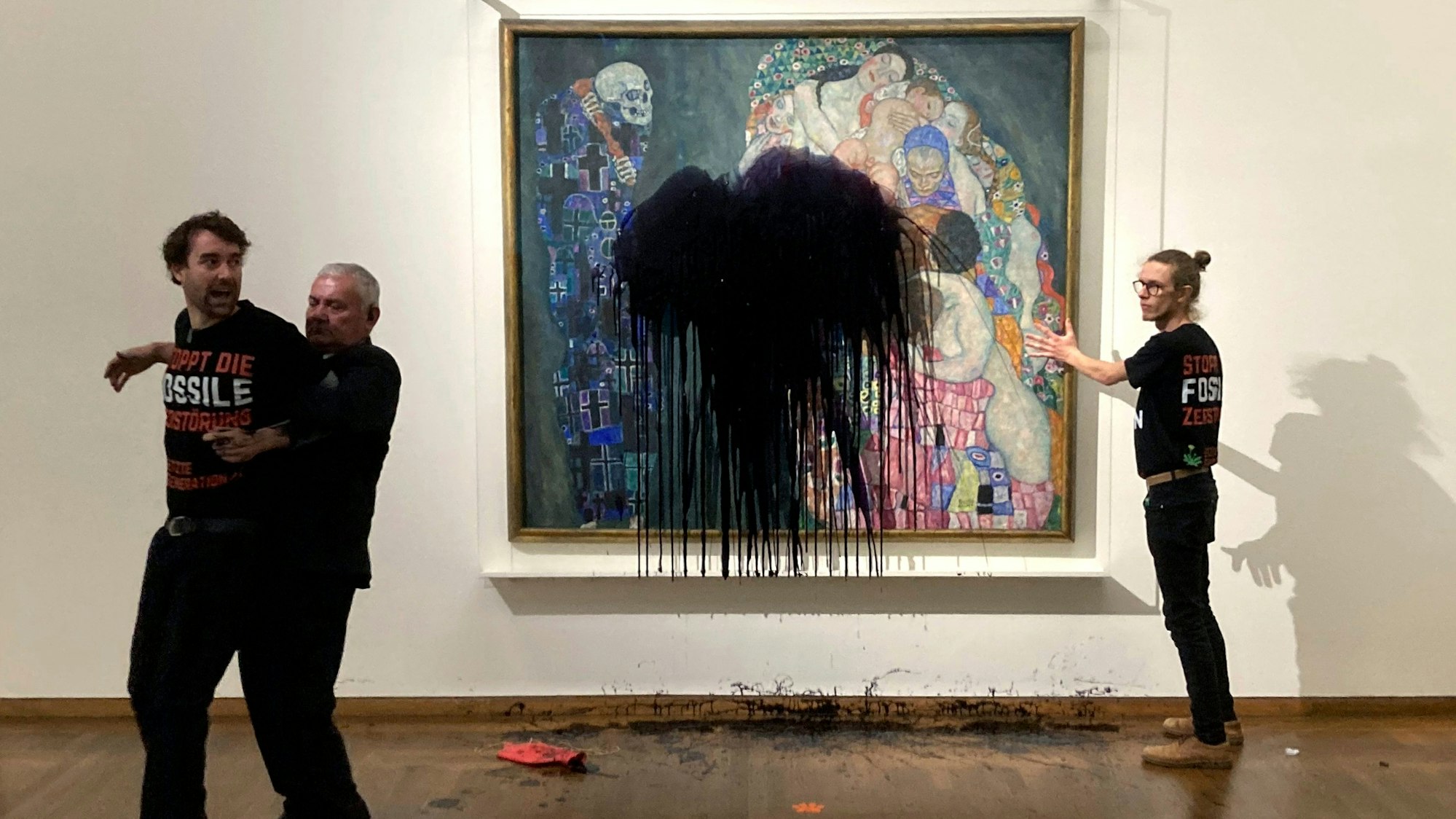 Ein Sicherheitsmann trägt einen Klima-Aktivisten von einem Klimt-Gemälde weg, auf das zuvor Öl geworfen wurde. Ein zweiter Aktivist steht neben dem Bild, auf dem ein großer Ölfleck klafft, das Bild wurde durch eine Glasscheibe geschützt.
