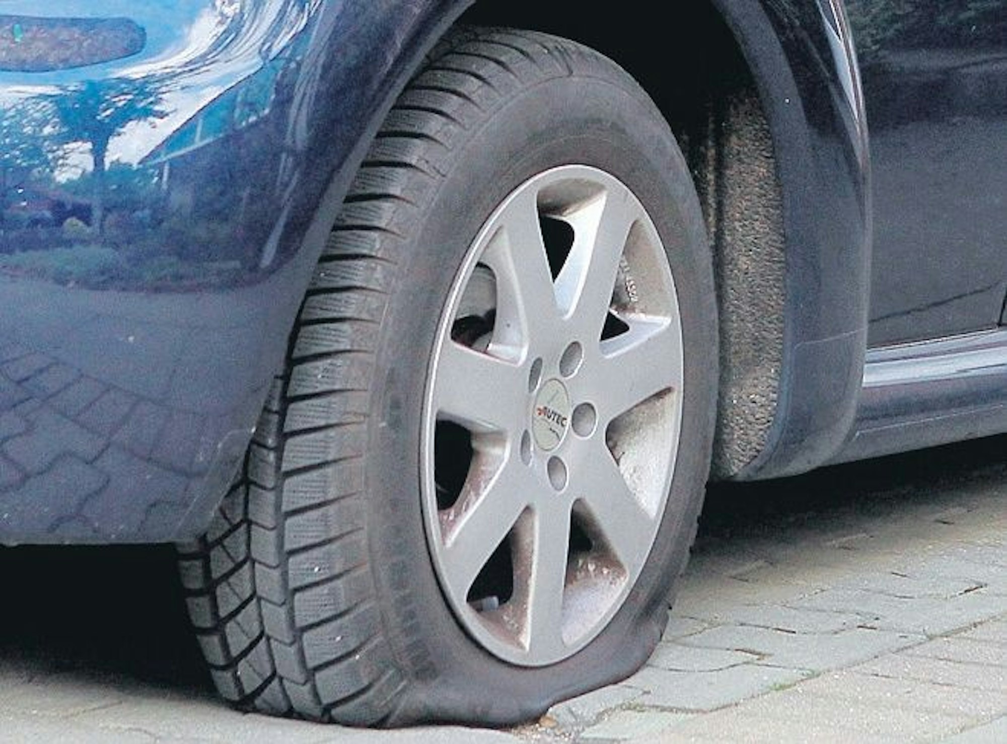 Ein blaues Auto mit einem platten Reifen steht auf Kopfsteinpflaster.