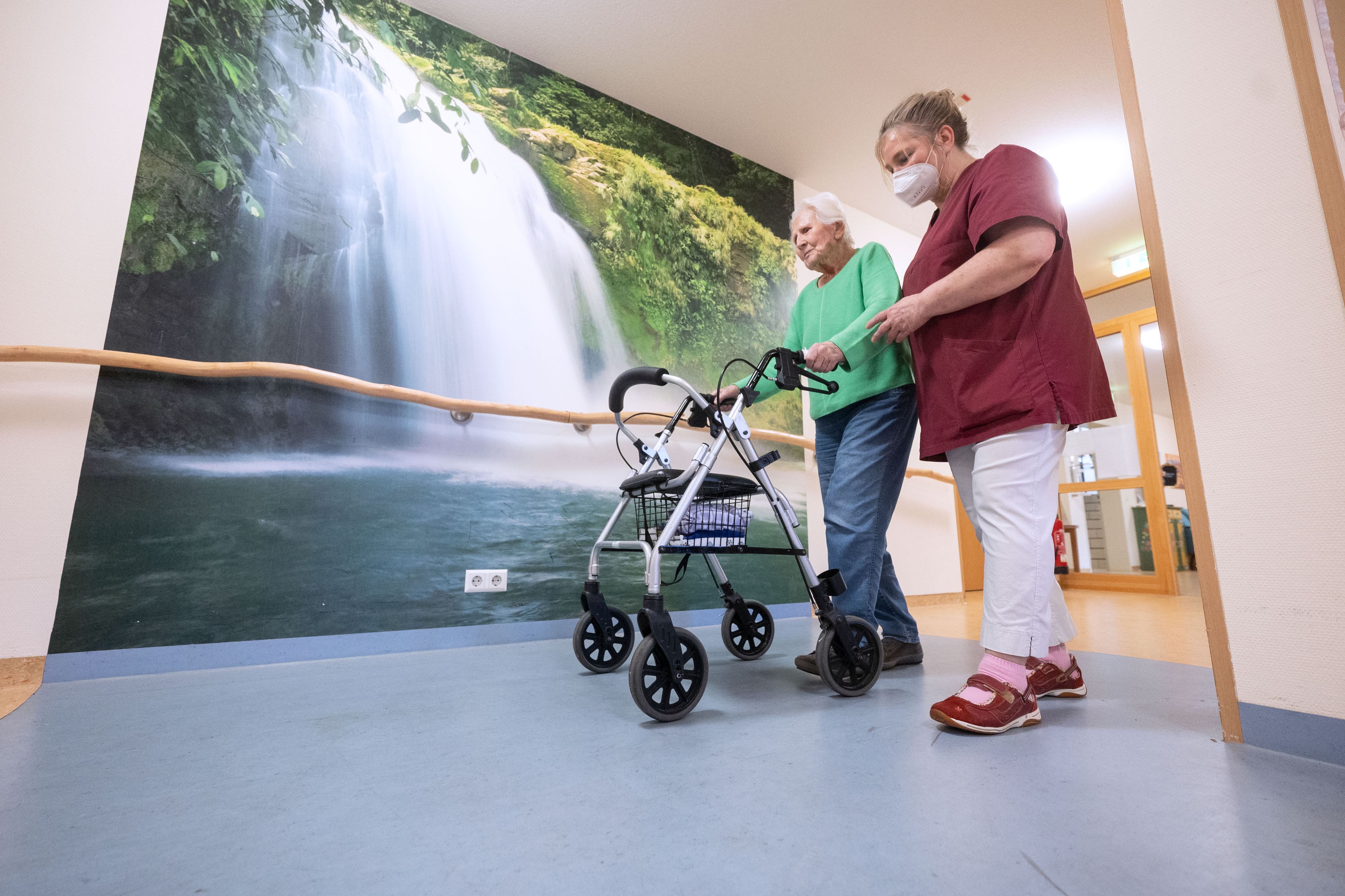 Das Foto zeigt einen Mann, der einen Rollator schiebt, mit einer Pflegeperson