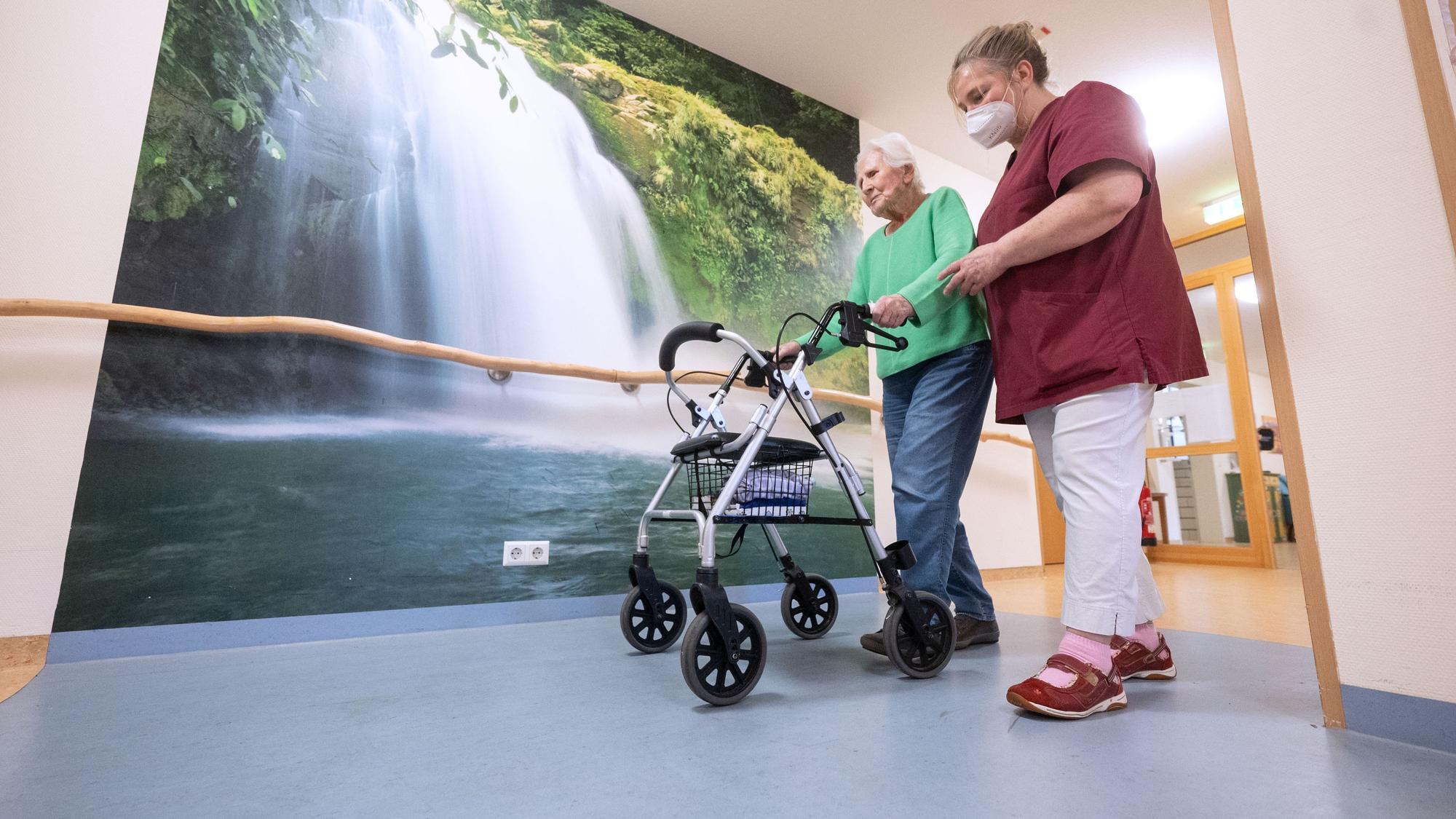 Das Foto zeigt einen Mann, der einen Rollator schiebt, mit einer Pflegeperson