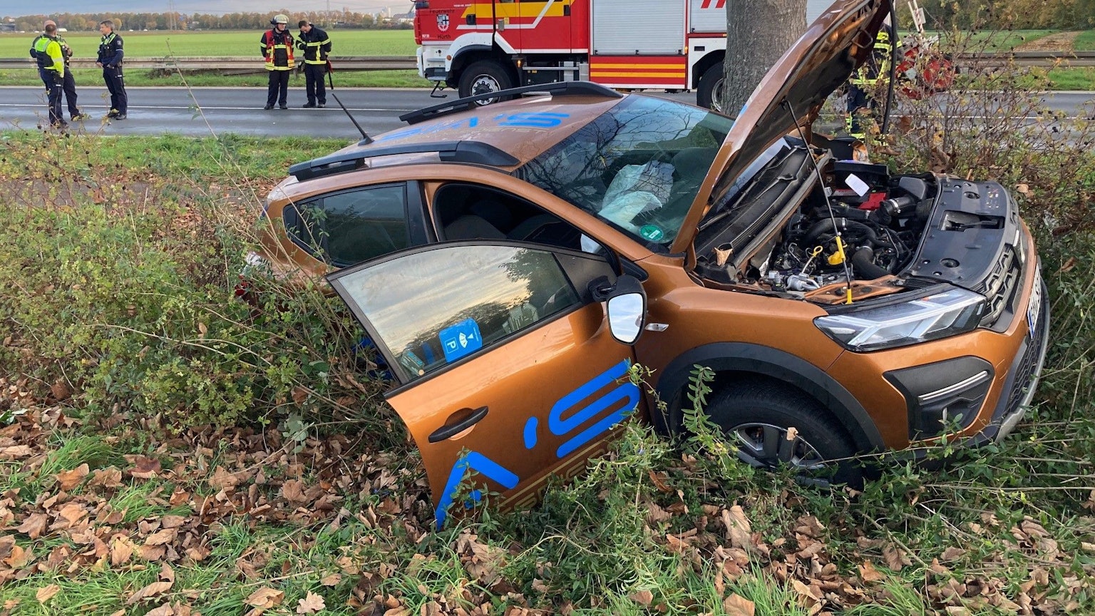 Ein Auto liegt im Straßengraben. Die Motorhaube und die Beifahrertür sind geöffnet. Auf der Straße im Hintergrund steht ein Feuerwehrauto, Polizisten und Feuerwehrleute.