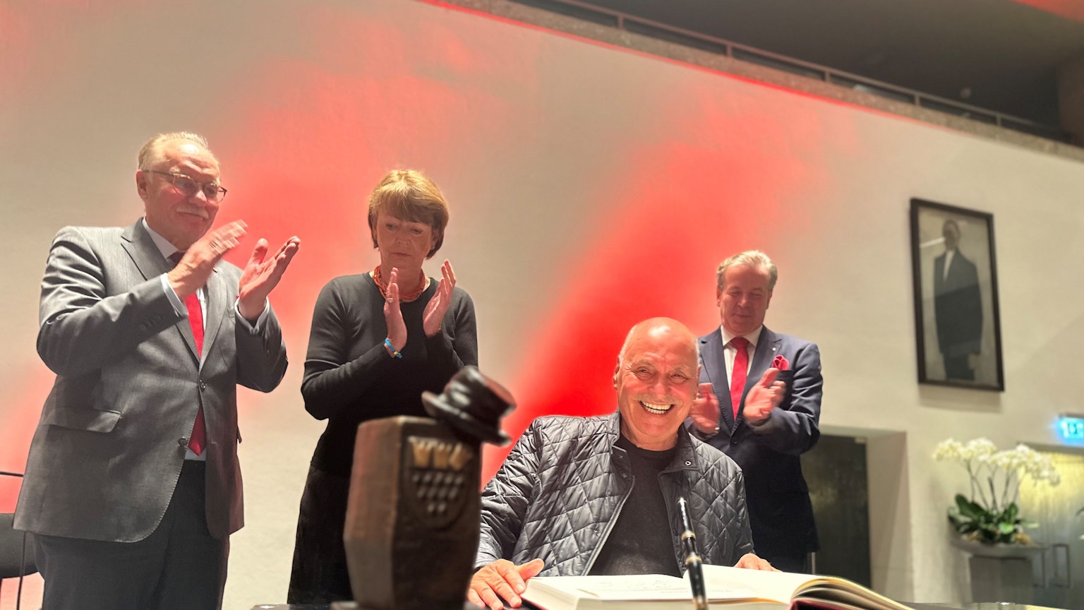 Rolly Brings wird mit dem Karl-Küpper-Preis ausgezeichnet und trägt sich ins Goldene Buch der Stadt Köln ein. Im Hintergrund applaudieren OB Henriette Reker, Festkomitee-Präsident Christoph Kuckelkorn und Bernhard Conin.