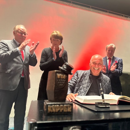 Rolly Brings wird mit dem Karl-Küpper-Preis ausgezeichnet und trägt sich ins Goldene Buch der Stadt Köln ein. Im Hintergrund applaudieren OB Henriette Reker, Festkomitee-Präsident Christoph Kuckelkorn und Bernhard Conin.