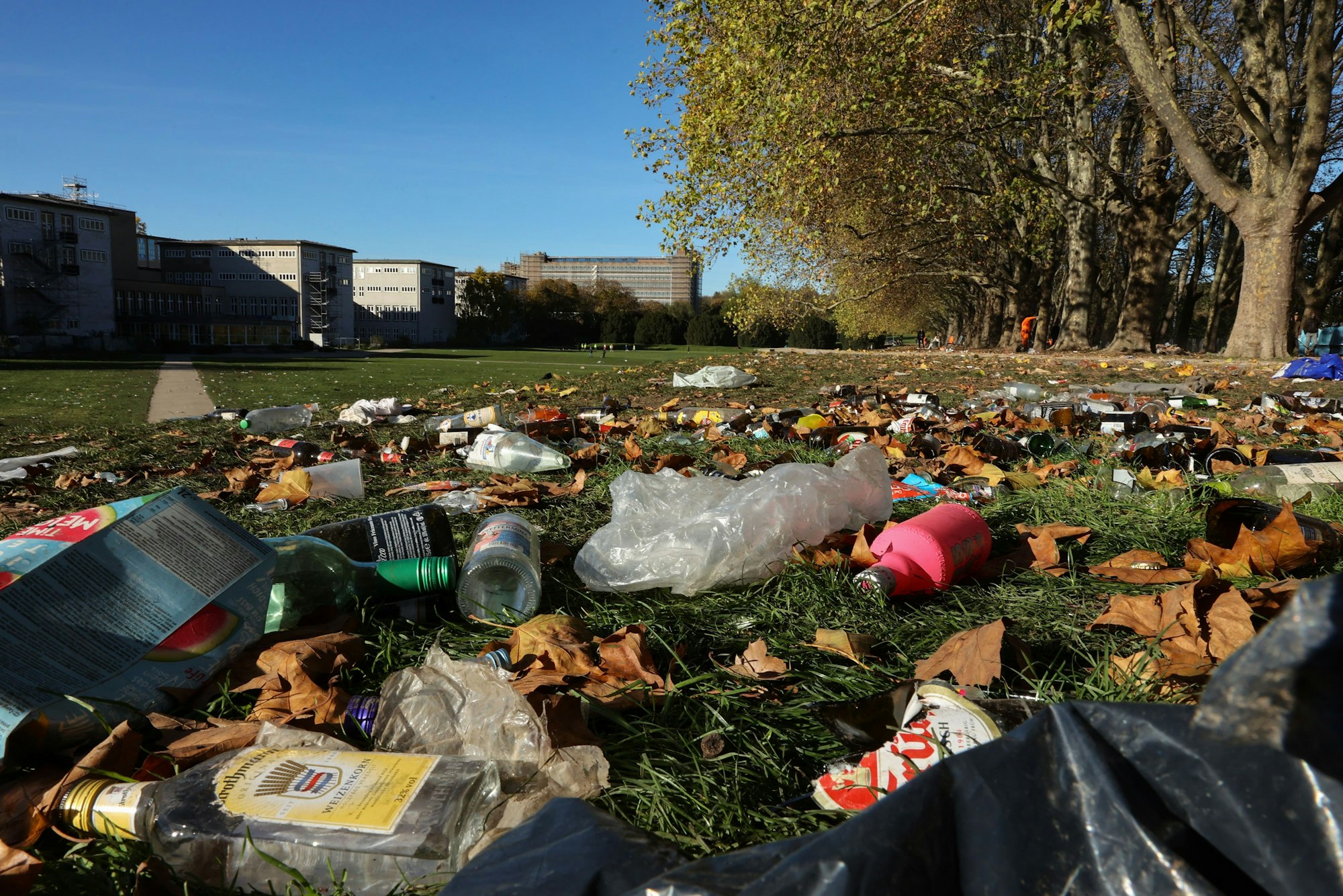 Blick auf eine große Wiese, die rechts von Bäumen, links von Unigebäuden gesäumt wird und auf der Mengen von Glasflaschen und Plastikmüll liegen