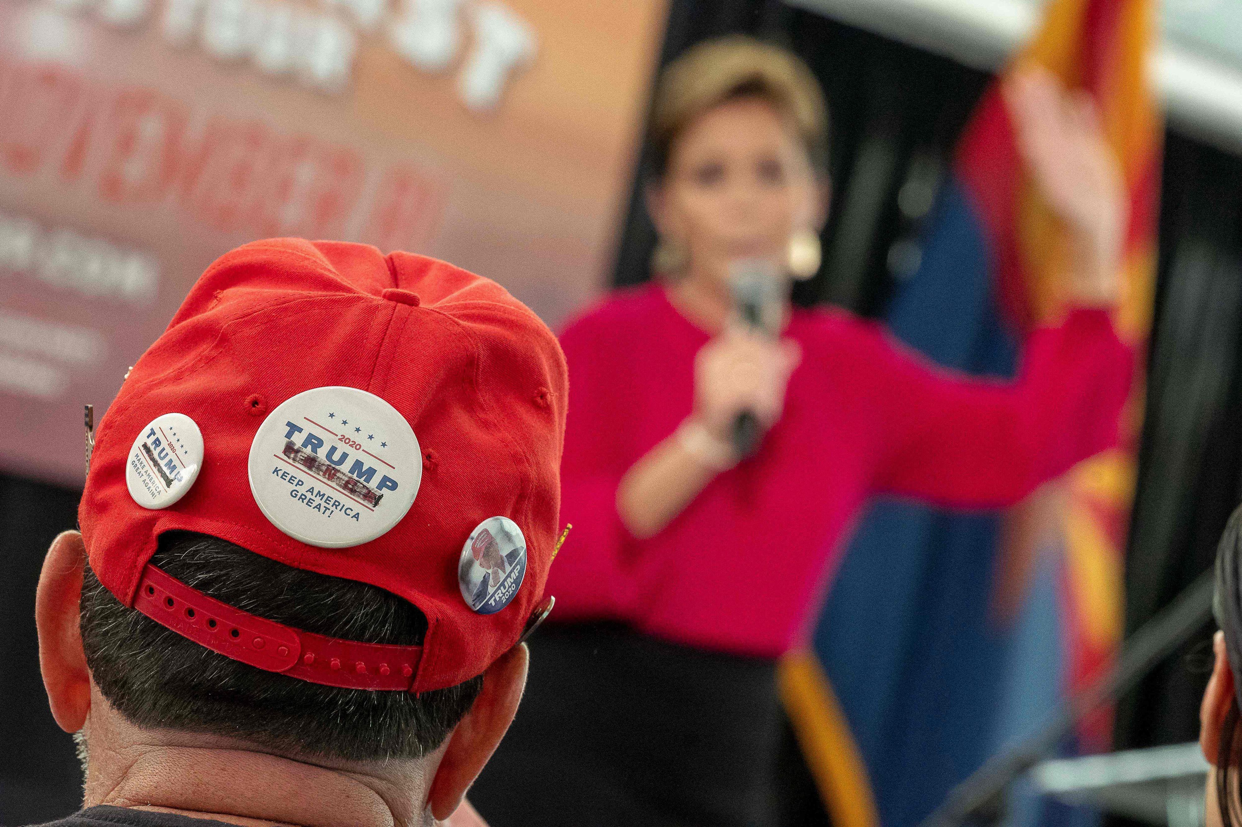 Im Vordergrund kann man einen Mann mit einer Kappe sehen, auf der ein Trump-Button ist. Im Hintergrund ist Kari Lake zu sehen.