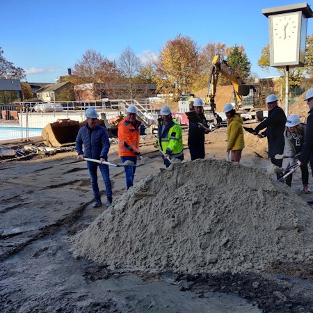 Vertreter der Stadt und der Bauunternehmen stechen auf der Baustelle im Freibad mit Schaufeln in einen Sandhaufen.