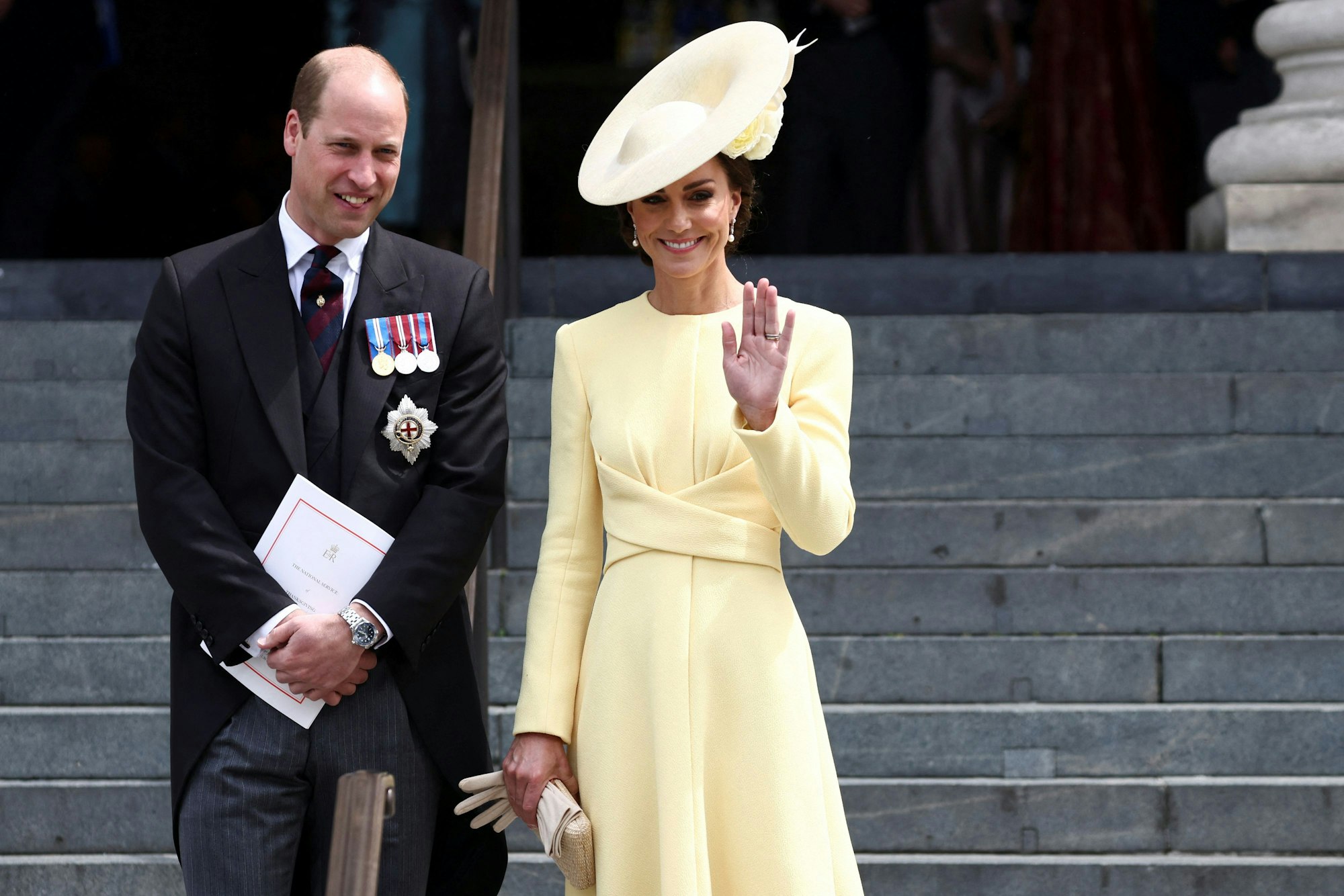 Prinz William und Herzogin Kate stehen auf einer Treppe. Sie tragen festliche Kleidung und lächeln. William hat die Hände verschränkt, Kate winkt in die Kamera.