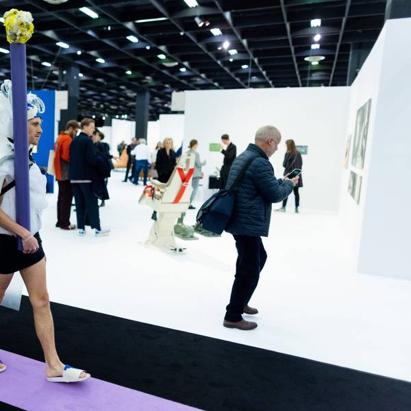 Ein Performance-Künstler mit weißer Schleppe und Pfosten geht durch eine Messehalle der Kunstmesse Art Cologne.