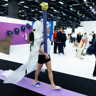 Ein Performance-Künstler mit weißer Schleppe und Pfosten geht durch eine Messehalle der Kunstmesse Art Cologne.