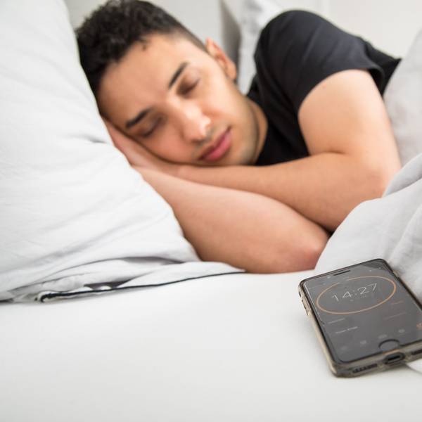 Ein junger Mann schläft im Bett, im Vordergrund ist sein Smartphone mit gestelltem Timer zu sehen.