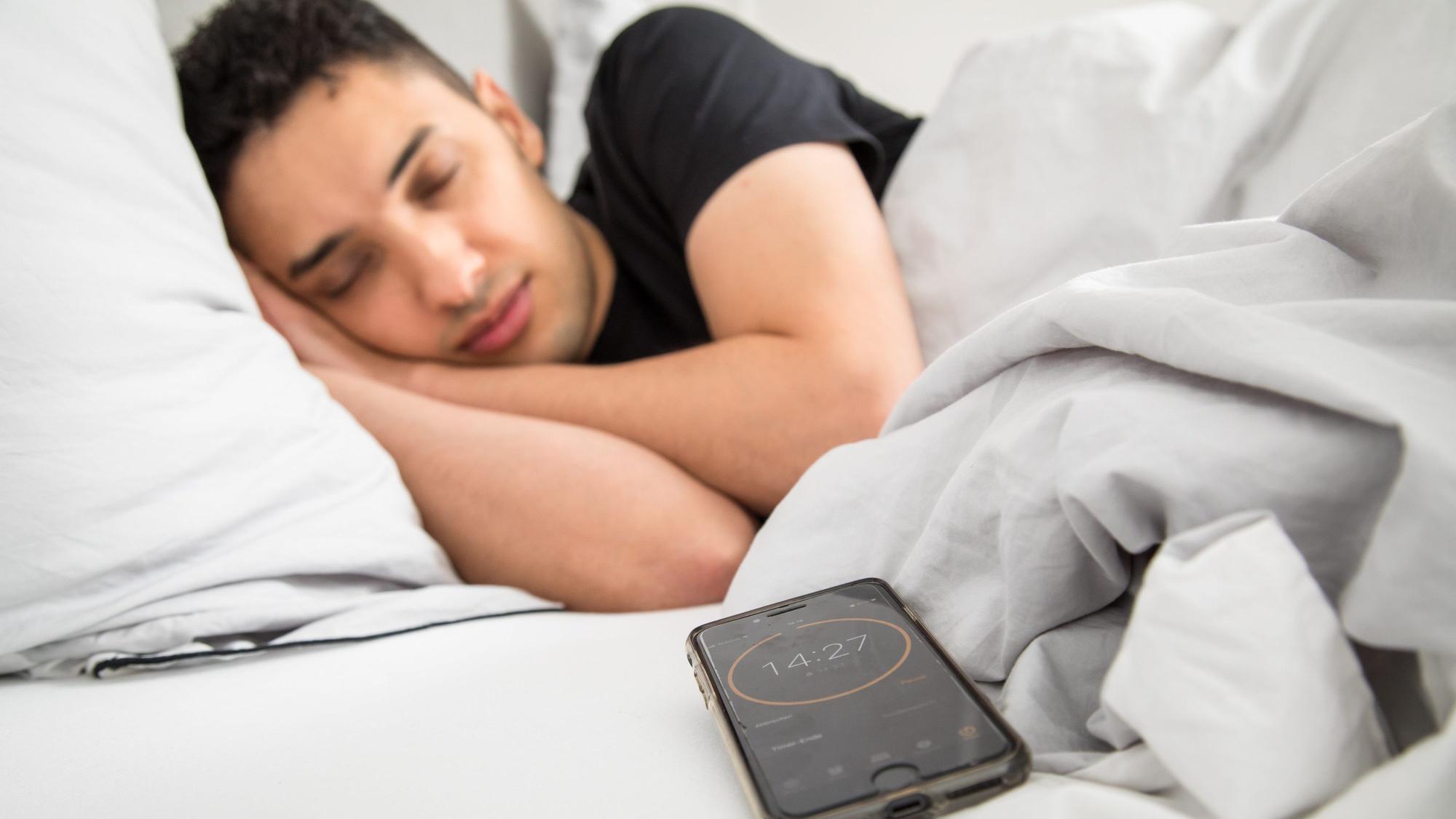 Ein junger Mann schläft im Bett, im Vordergrund ist sein Smartphone mit gestelltem Timer zu sehen.
