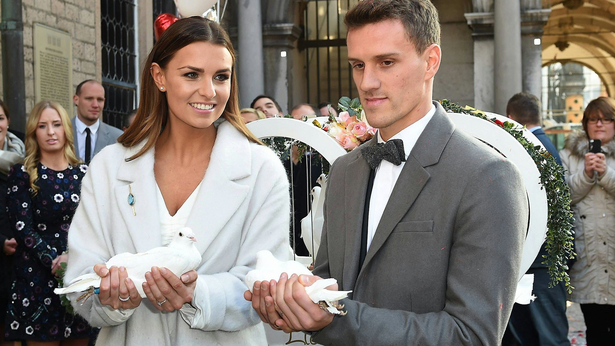 Simon Zoller und Laura Wontorra mit weißen Tauben vor dem Kölner Rathaus. Sie lächeln und tragen Hochzeitskleidung. Sie in Weiß, er in grauem Anzug.
