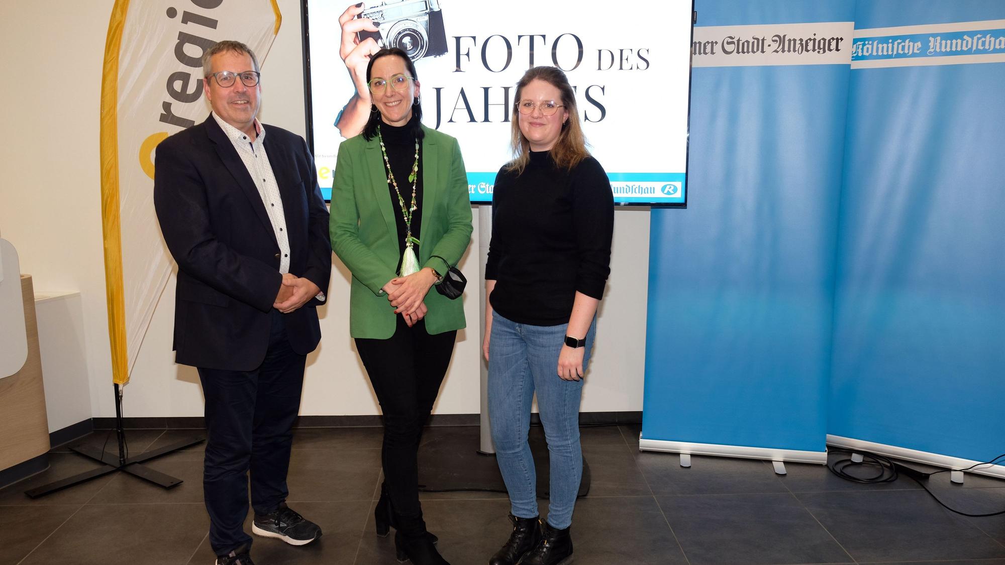 Ein Mann und zwei Frauen stehen vor einem Bildschirm mit der Aufschrift „Foto des Jahres“. Daneben stehen Banner vom „Kölner Stadt-Anzeiger“ und „Kölnische Rundschau“.